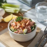 Close-up overhead view of a creamy tuna salad with eggs garnished with parsley and paprika, served in a white bowl with lemon wedges and celery sticks on a wooden board.